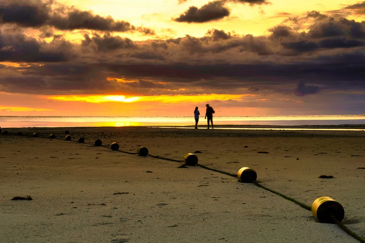 Rope lined with buoys marking the swimming area on Tamarin beach at sunset, with two silhouetted people near the sea.