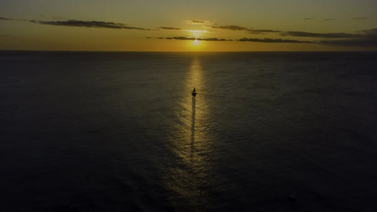 A sailboat aligned with the sunset reflection on the ocean in Mauritius, seen from an aerial perspective