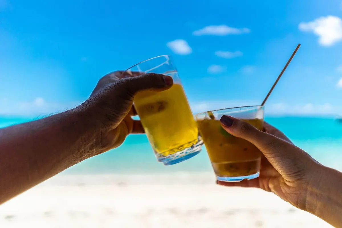 Two hands holding beer and cocktail glasses cheering on a Mauritius beach