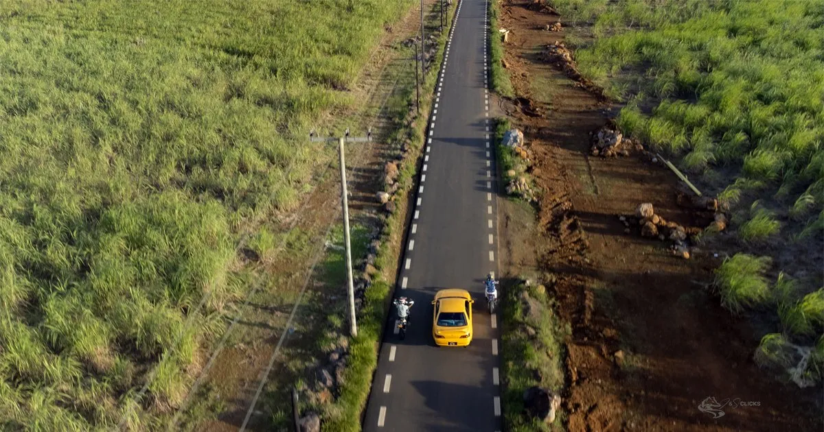 Aerial view of a yellow car driving on the road to Mare Longue between sugarcane fields and green rural landscapes.
