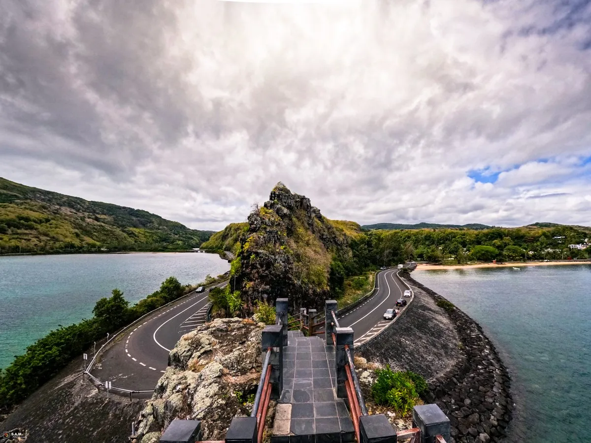 View from the stairs at Macondé showing the curved coastal road, rocky viewpoint, lagoon, and green hills.