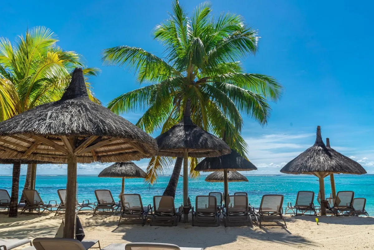 Thatched umbrellas and beach chairs on Le Paradis Beachcomber shoreline in Mauritius with turquoise ocean background