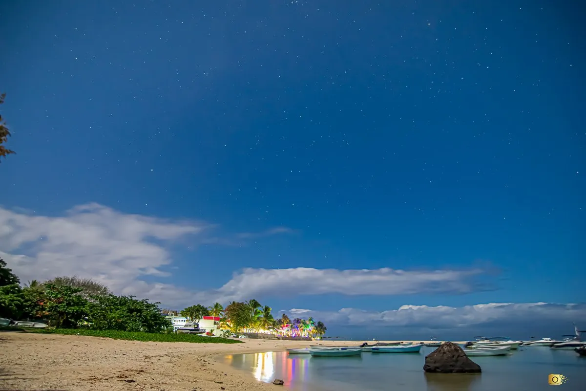 Night view of Flic en Flac beach with stars, anchored boats, calm lagoon water, and colourful lights from Villa Caroline Hotel.