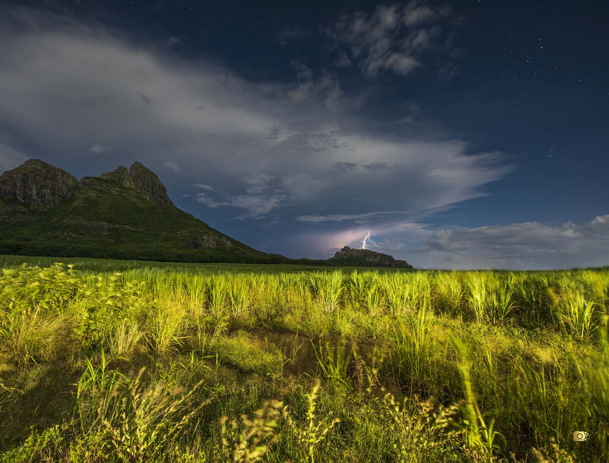 Lightning strike over a sugarcane field in Mauritius at night, with illuminated clouds and distant mountains in the background.