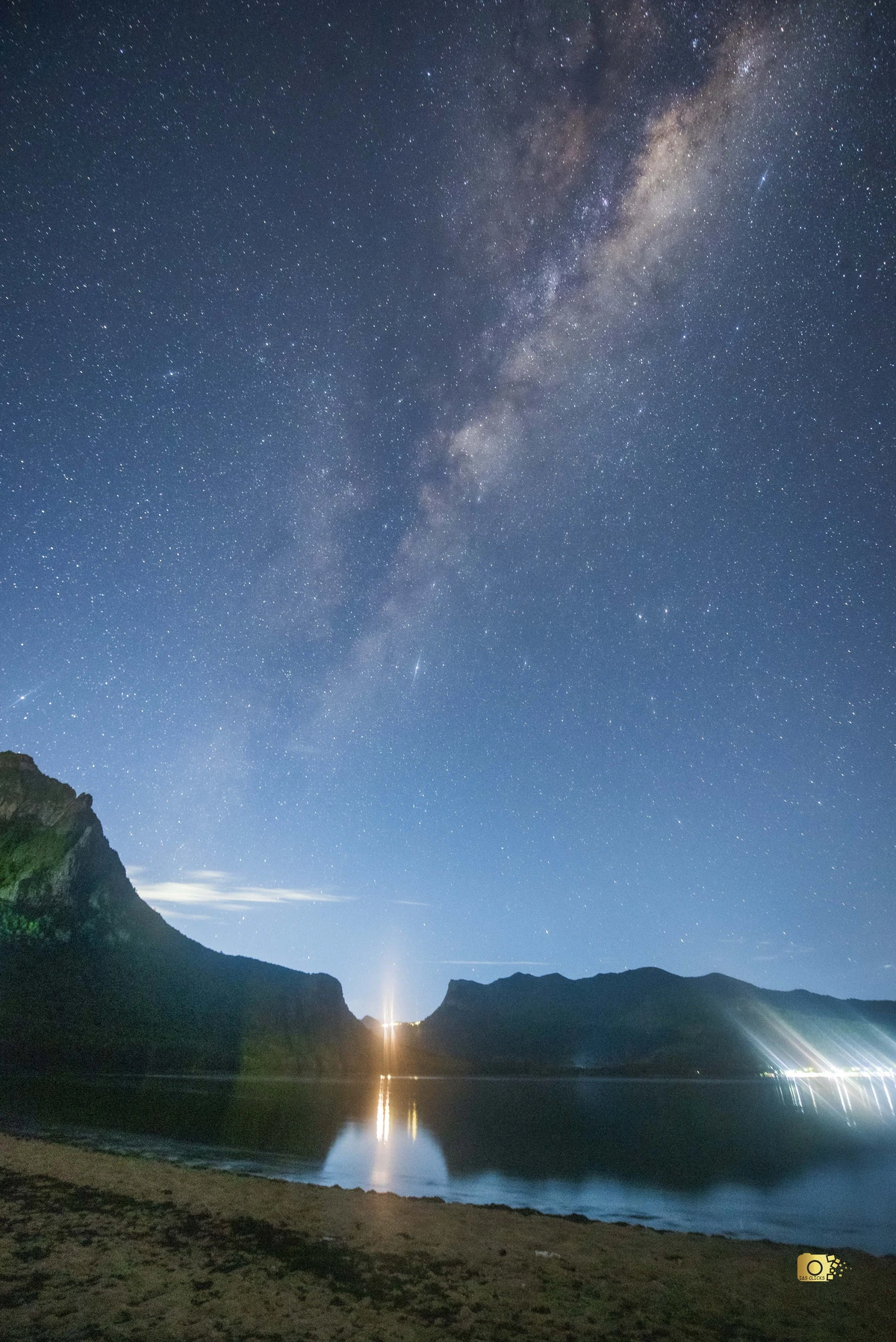 Milky Way above Le Morne lagoon at night, with part of Le Morne mountain, calm water reflections, and beach lights.
