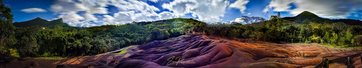 Panoramic view of the Seven Coloured Earths in Chamarel, Mauritius, with colourful dunes, surrounding forest, and dramatic clouds at night.