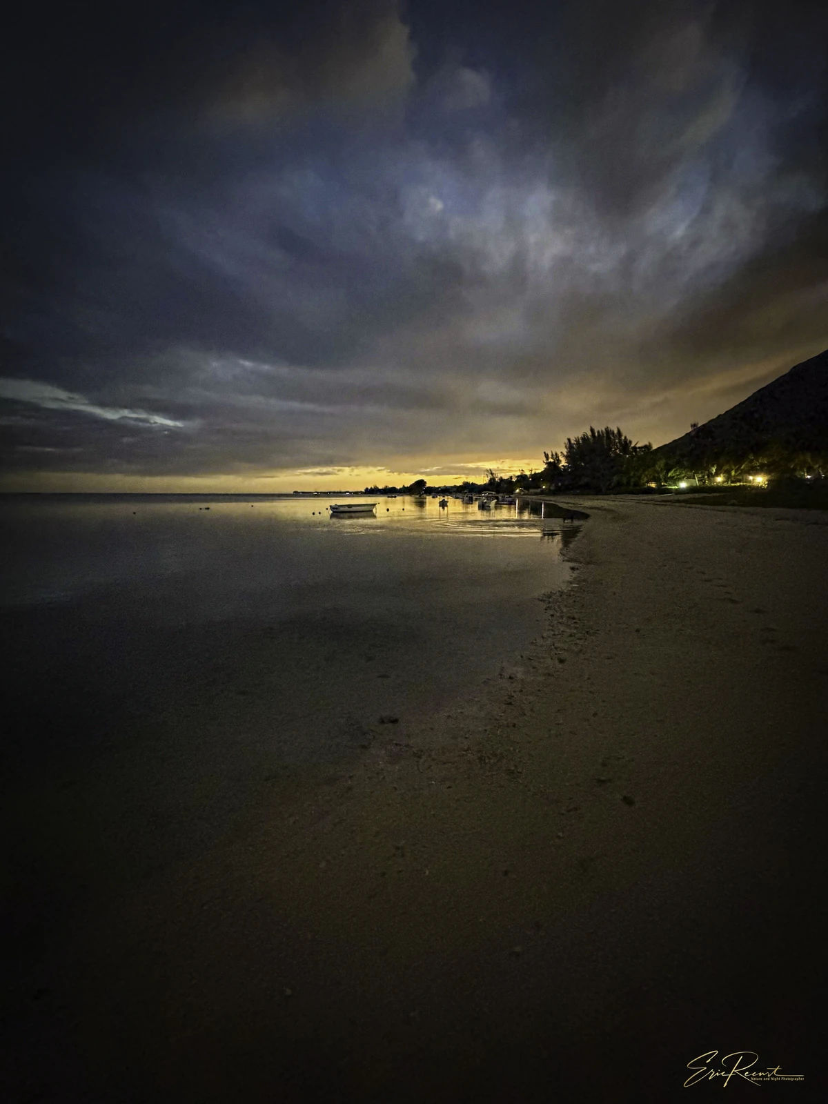 Early morning view of La Prairie beach in Mauritius with calm lagoon water, dark shoreline, distant lights, and dramatic clouds.