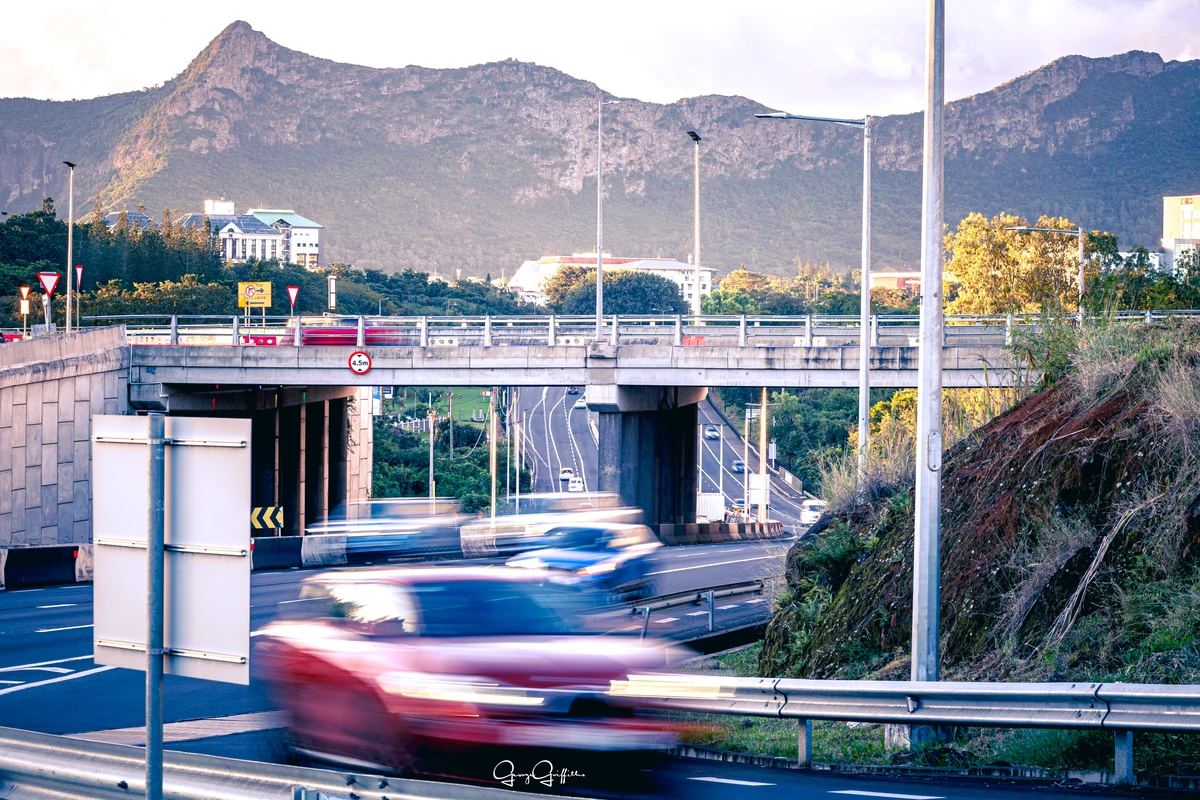 View of the Moka Range and Réduit from Tribeca Mall, with blurred traffic, road bridges, and mountains in the background.