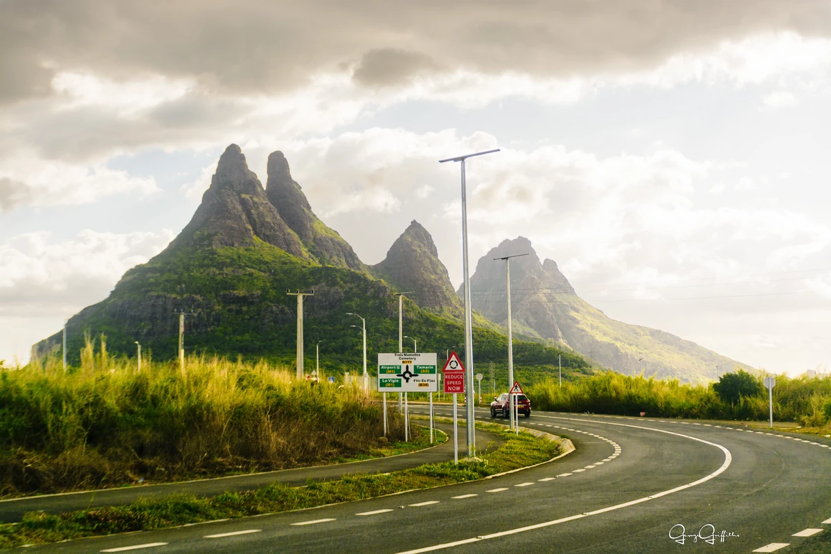 View of Trois Mamelles from the new La Vigie to Cascavelle road corridor, with cars, road signs, sugarcane fields, and mountains in Mauritius.