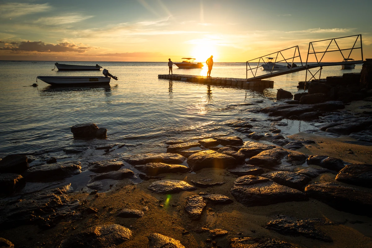 Sunset at Le Canonnier Hotel beach in Pointe aux Canonniers, with two silhouetted people on a jetty, flat rocks on the beach, and boats on the sea.