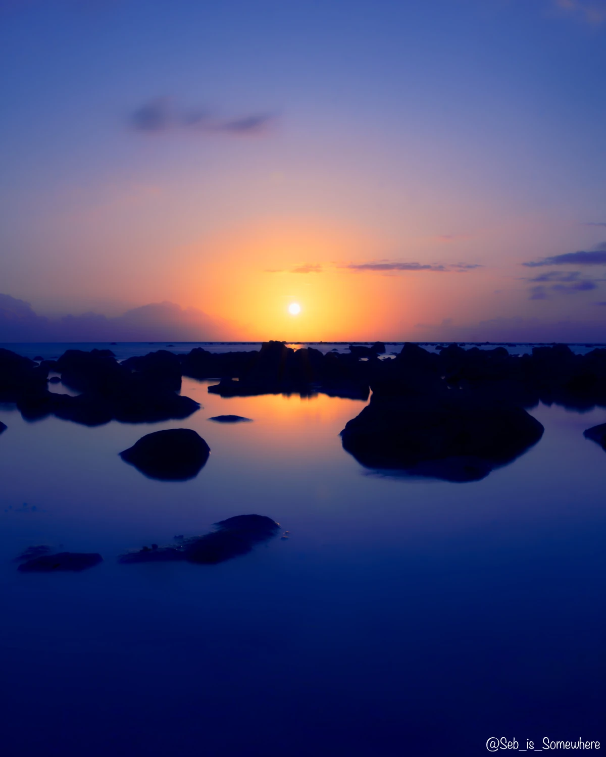 Long-exposure sunset at a rocky part of Albion beach in Mauritius, with dark rocks, calm water, and soft golden reflections.