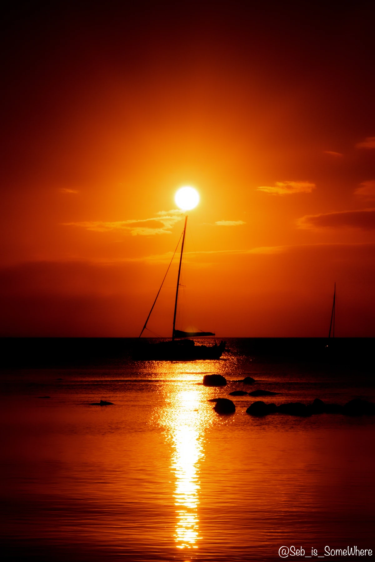 Sailboat silhouette at sunset in Mont Choisy, Mauritius, with an intense orange sky, golden reflection, calm water, and rocky shapes in the foreground.