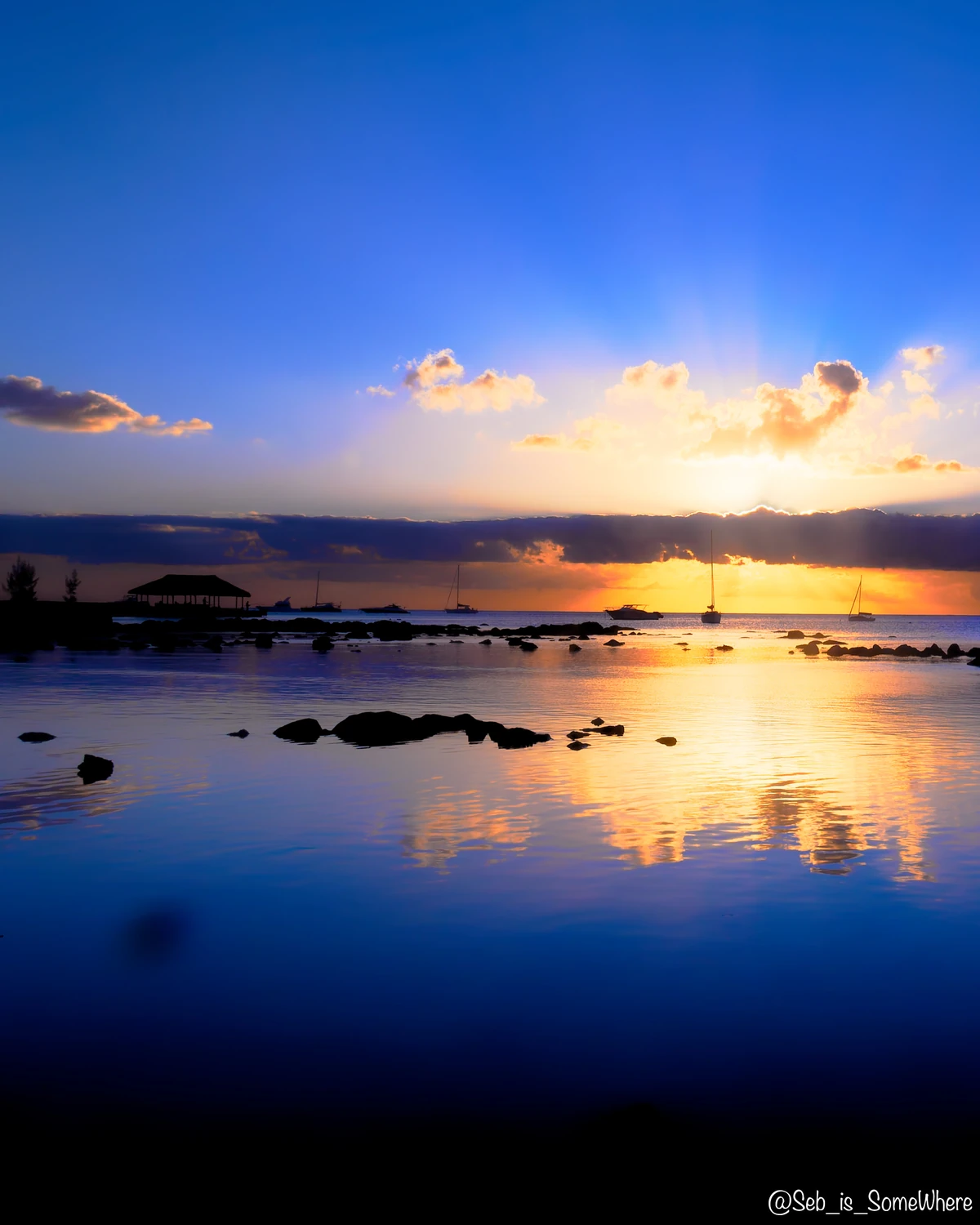 Crepuscular rays over Mont Choisy beach at sunset, with calm reflective water, silhouetted rocks, boats, and a thatched pavilion.