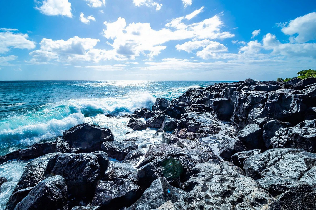 Waves crashing against dark volcanic rocks in front of Club Med Albion, Mauritius, under a bright blue sky.