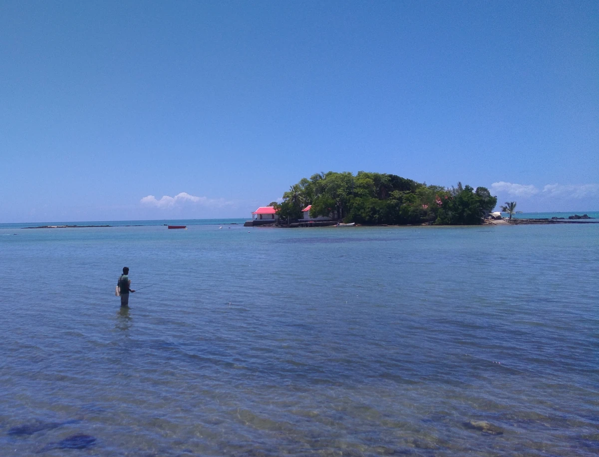 Mauritian fisherman standing in the shallow lagoon of Mahébourg with Île du Mouchoir Rouge in the background.