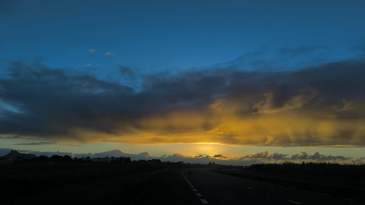 Sunrise over a road in Plaine Magnien, Mauritius, with dramatic clouds, golden light, and an empty road leading toward the horizon.