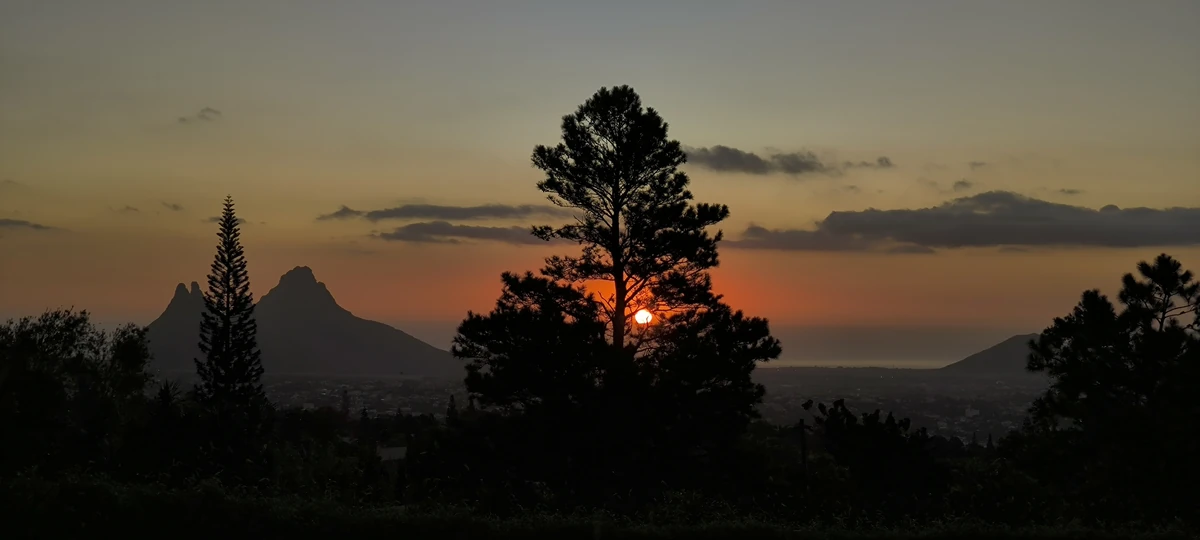 Sunset view of Trois Mamelles from Trou aux Cerfs in Curepipe with silhouetted trees in the foreground.
