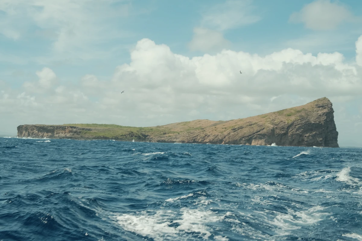 Close-up view of Coin de Mire from the sea, showing its rocky coastline, dry vegetation and waves off the north coast of Mauritius.