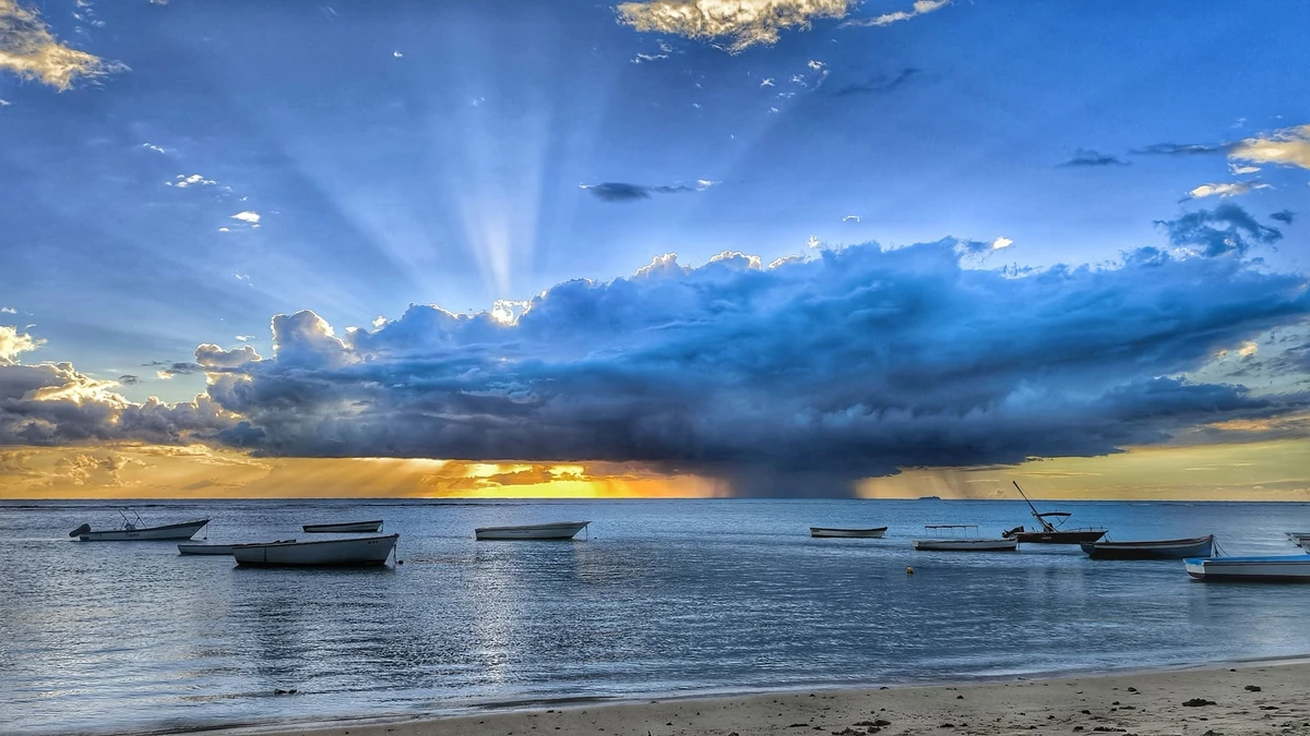 Crepuscular rays shining through clouds above Albion public beach, with small boats resting on calm sea water.