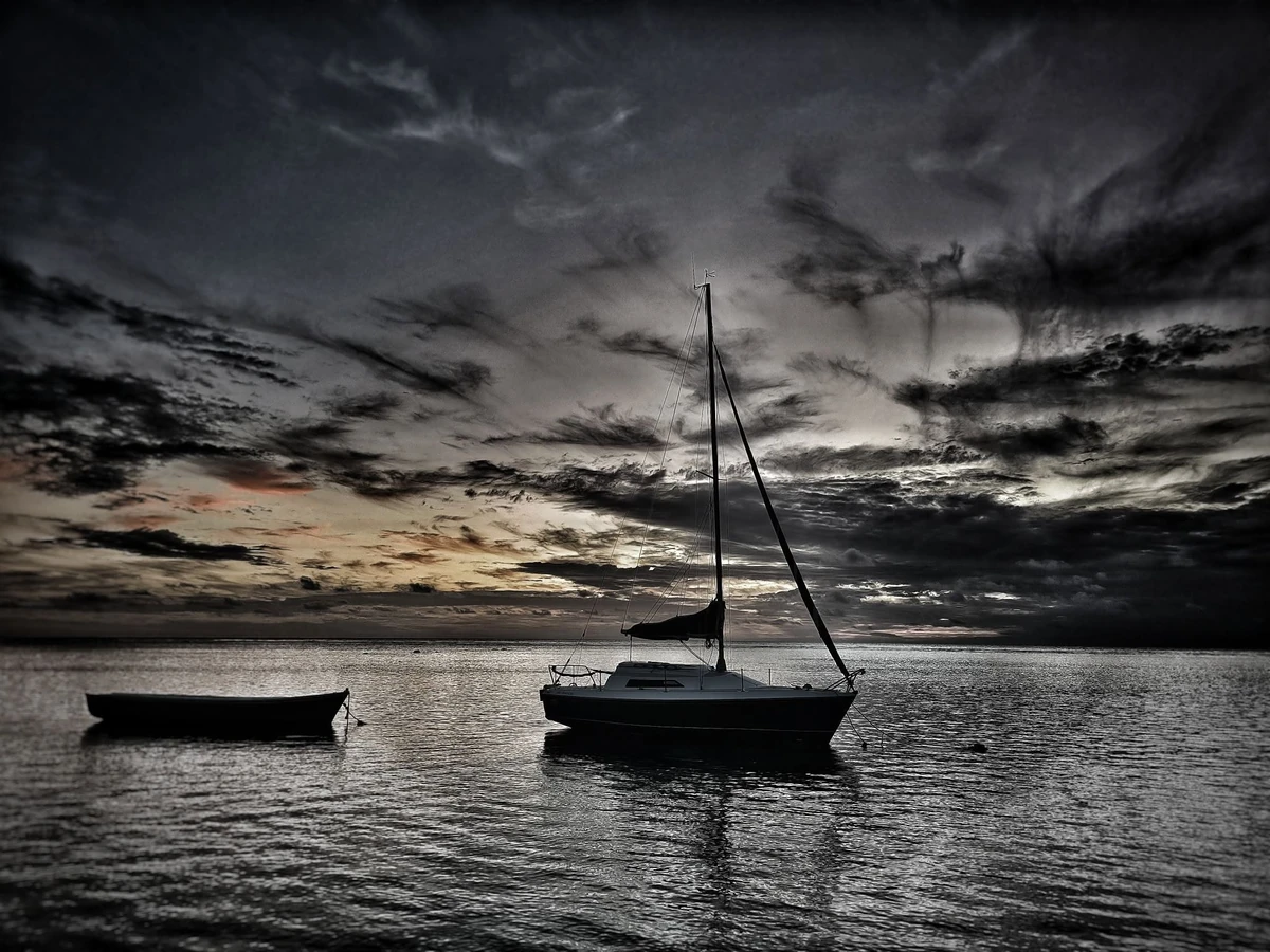 Sailboat and small boat in silhouette after sunset at Albion, Mauritius, with calm sea and dramatic dark clouds.