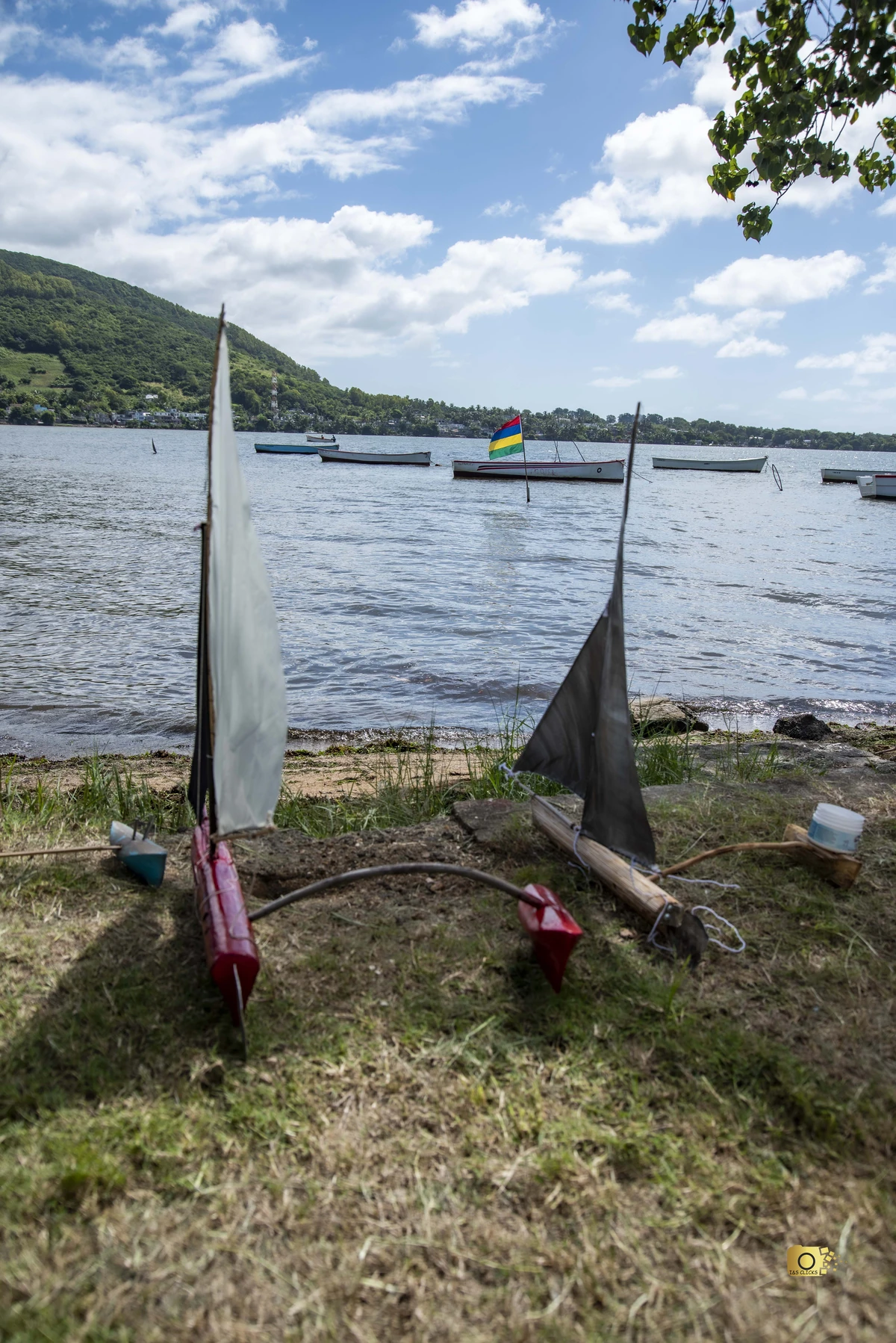 Small model sailing pirogues made of wood and raffia-like sails on the shore at Quatre Soeurs, with real boats in the lagoon behind them.