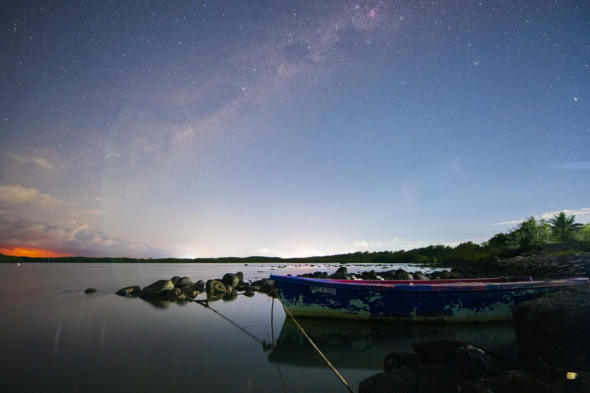 Fishing boat on calm water at Poudre d’Or under the Milky Way, with moonrise glow on the horizon.