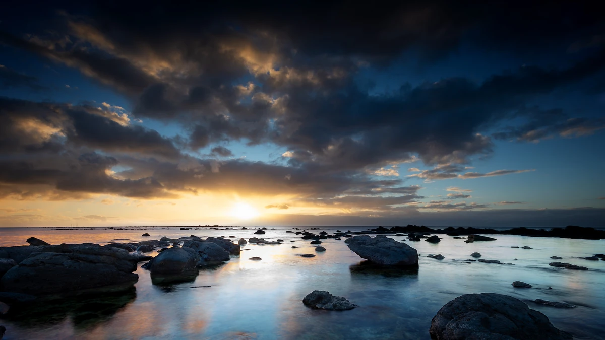 Dramatic sunset over the rocky shoreline of Flic en Flac in Mauritius, with scattered rocks in calm shallow water reflecting golden light beneath dark clouds