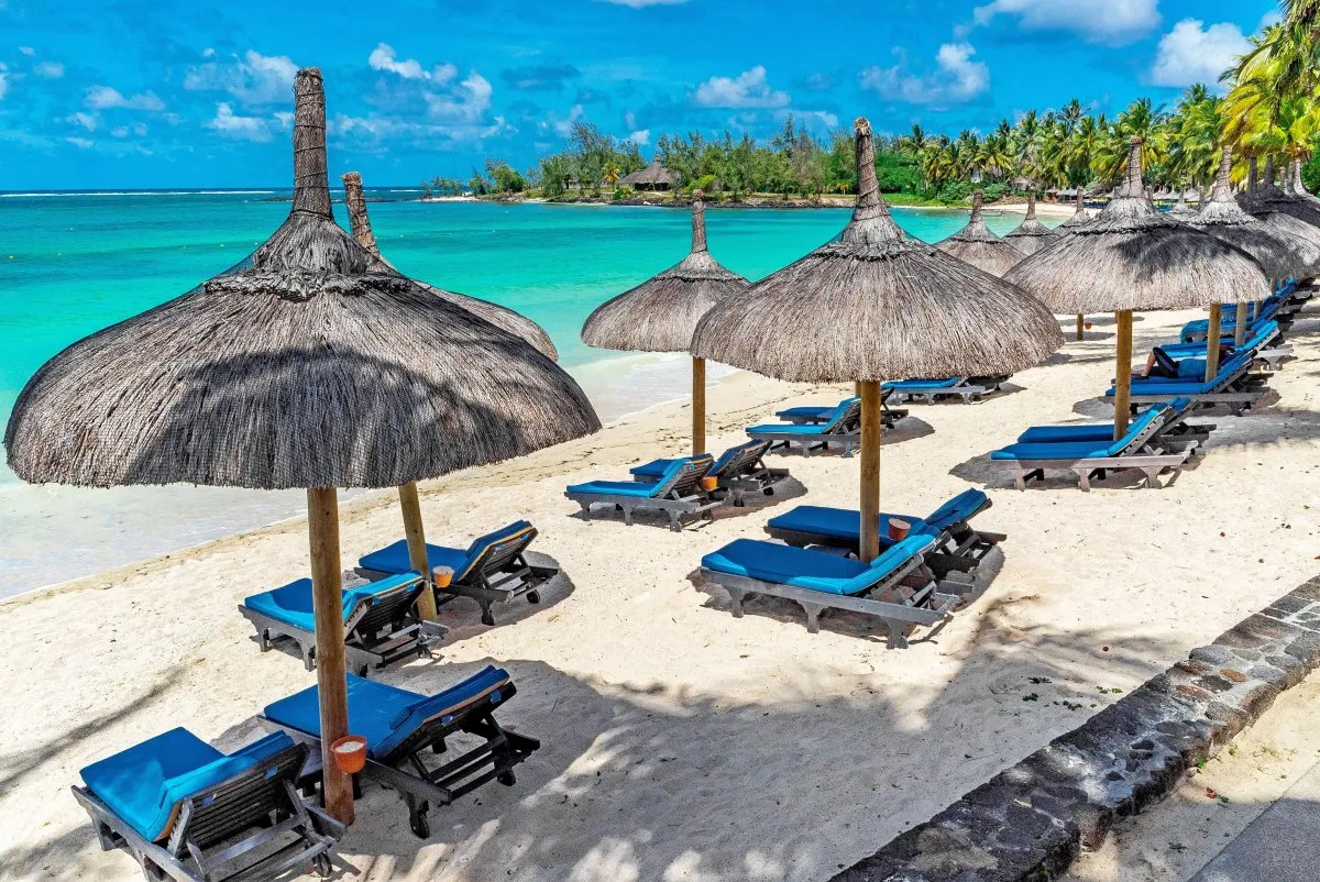 Thatched umbrellas and blue deckchairs on Constance Belle-Mare beach with palm trees and turquoise sea in Mauritius