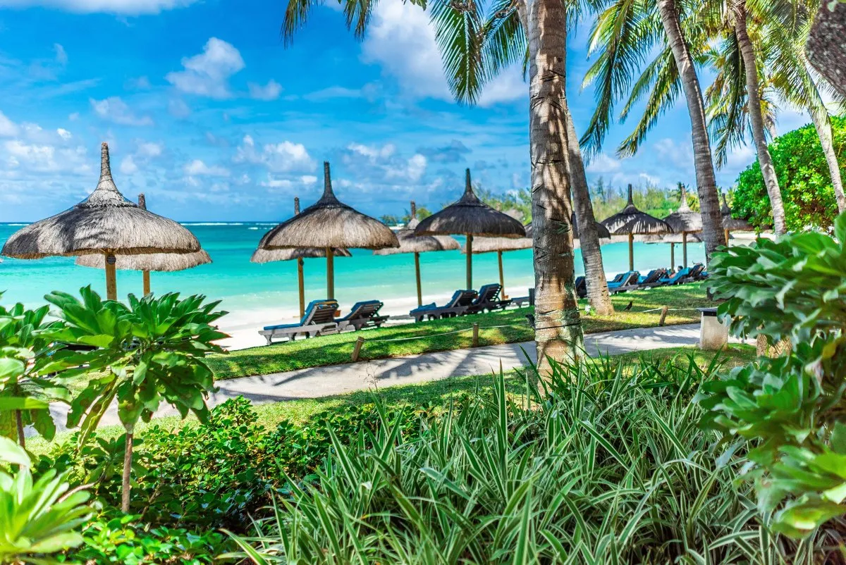 View of turquoise lagoon and thatched umbrella at Constance Belle-Mare beach in Mauritius with hotel and green plants