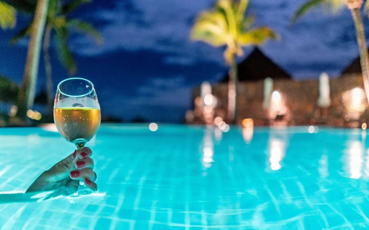 Hand holding wine glass by illuminated pool at Constance Belle-Mare Plage hotel in Mauritius at night