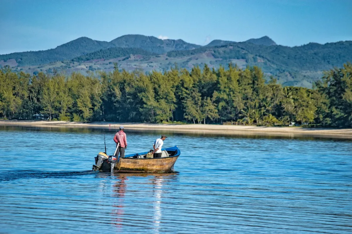 Fishermen in wooden boat setting out to fish at Riambel beach in Mauritius with trees and mountains nearby